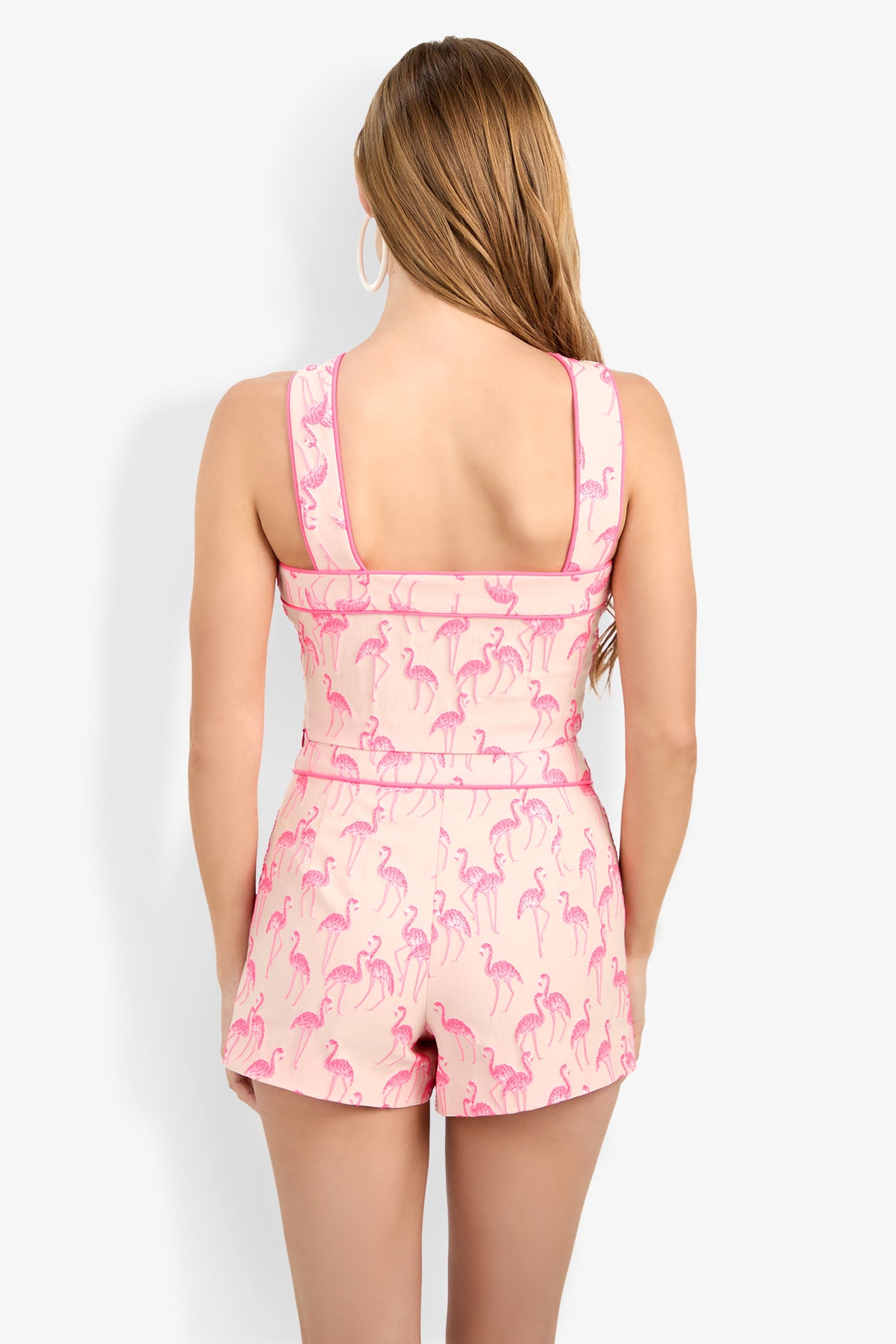 A woman with long, light brown hair is seen from the back wearing the sleeveless Piped Wanona Top in light pink with a flamingo pattern, large hoop earrings, and standing against a plain white background.