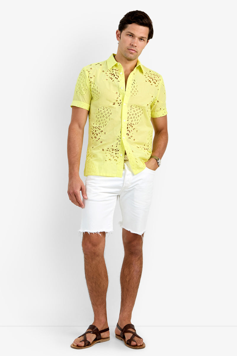 A man wearing the BRANSON SHIRT in Citrus, white frayed-hem 100% cotton shorts, brown sandals, and a wristwatch stands against a plain white background.