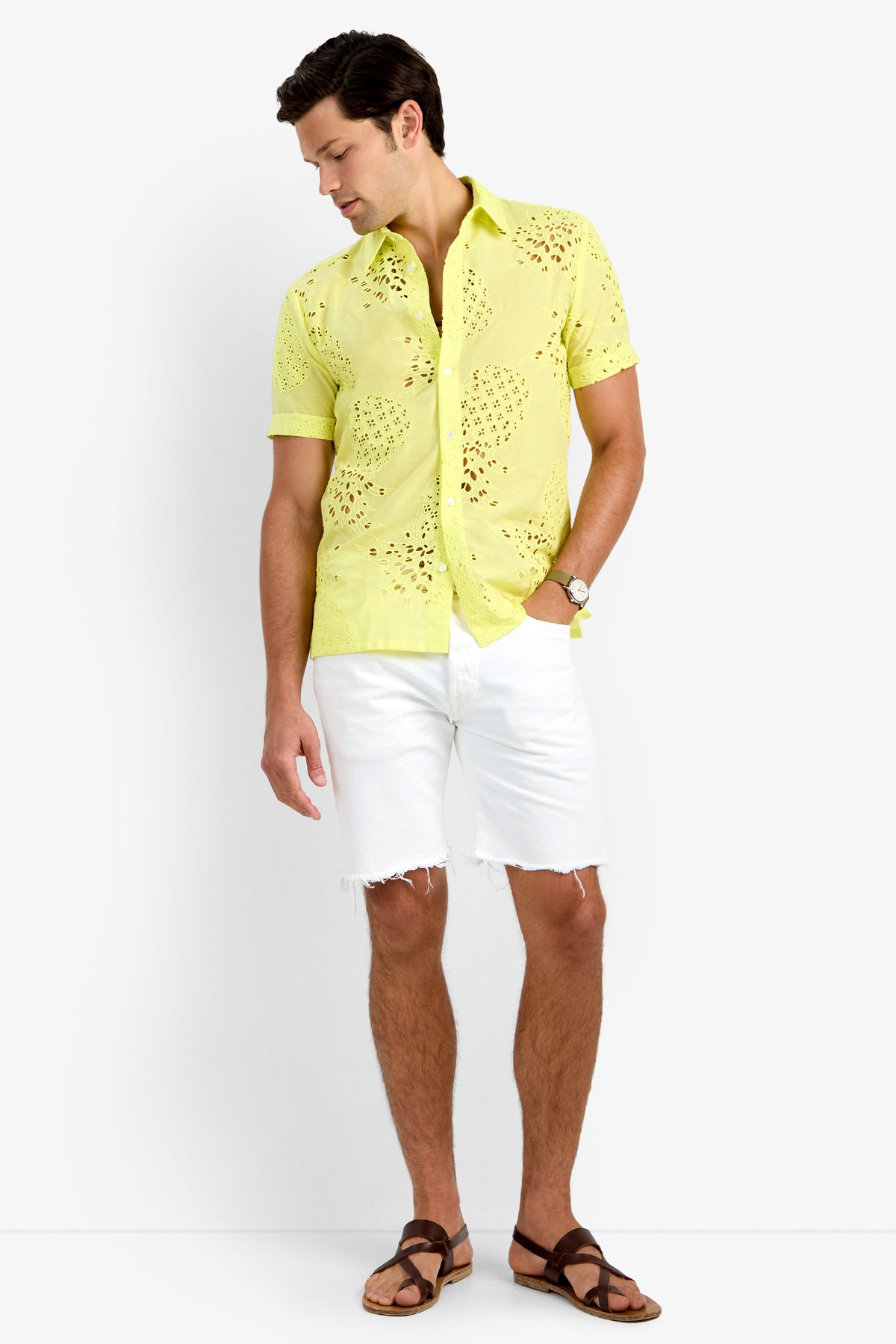 A man wearing the BRANSON SHIRT, a light yellow, short-sleeve 100% cotton eyelet button-up, paired with white frayed shorts and brown sandals, stands against a plain white background looking down to his left.