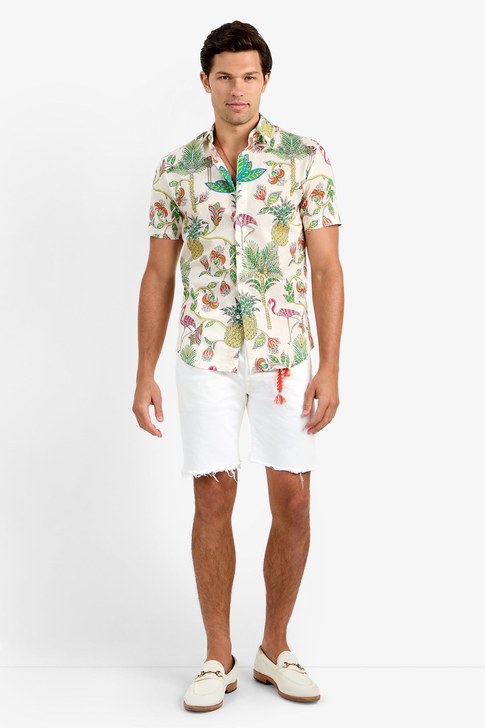 A man faces forward against a white background wearing the SLIM JIM SHIRT in Flamingo Fizz, a colorful tropical print 100% cotton short-sleeve shirt, paired with white frayed hem shorts and white loafers, looking relaxed and casual.