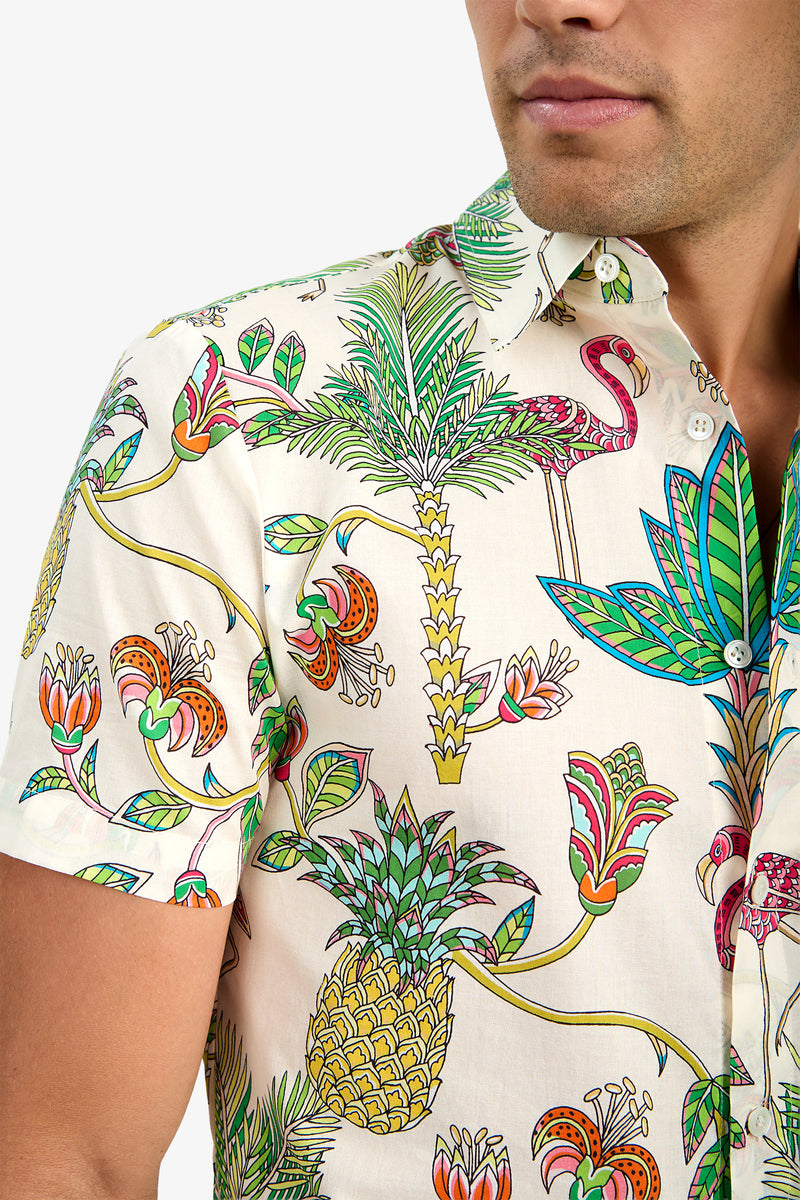A man wears the SLIM JIM SHIRT, a short-sleeved, 100% cotton button-up featuring a vibrant tropical print with pineapples, palm trees, flamingos, flowers, and leaves on a light background. Only his lower face and upper torso are visible.