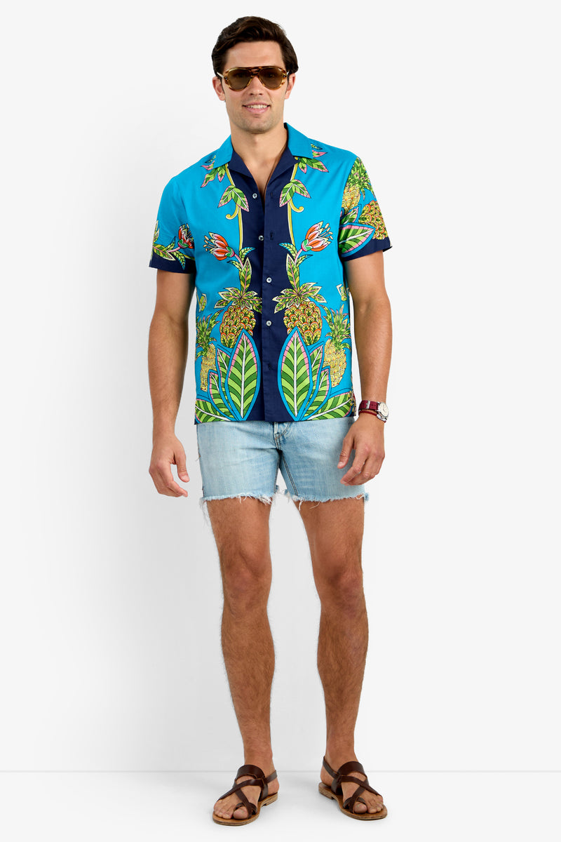 A man wearing sunglasses, a Tigerlily JOSUE CAMP SHIRT with a tropical pattern, light denim shorts, brown sandals, and a watch stands against a plain white background.