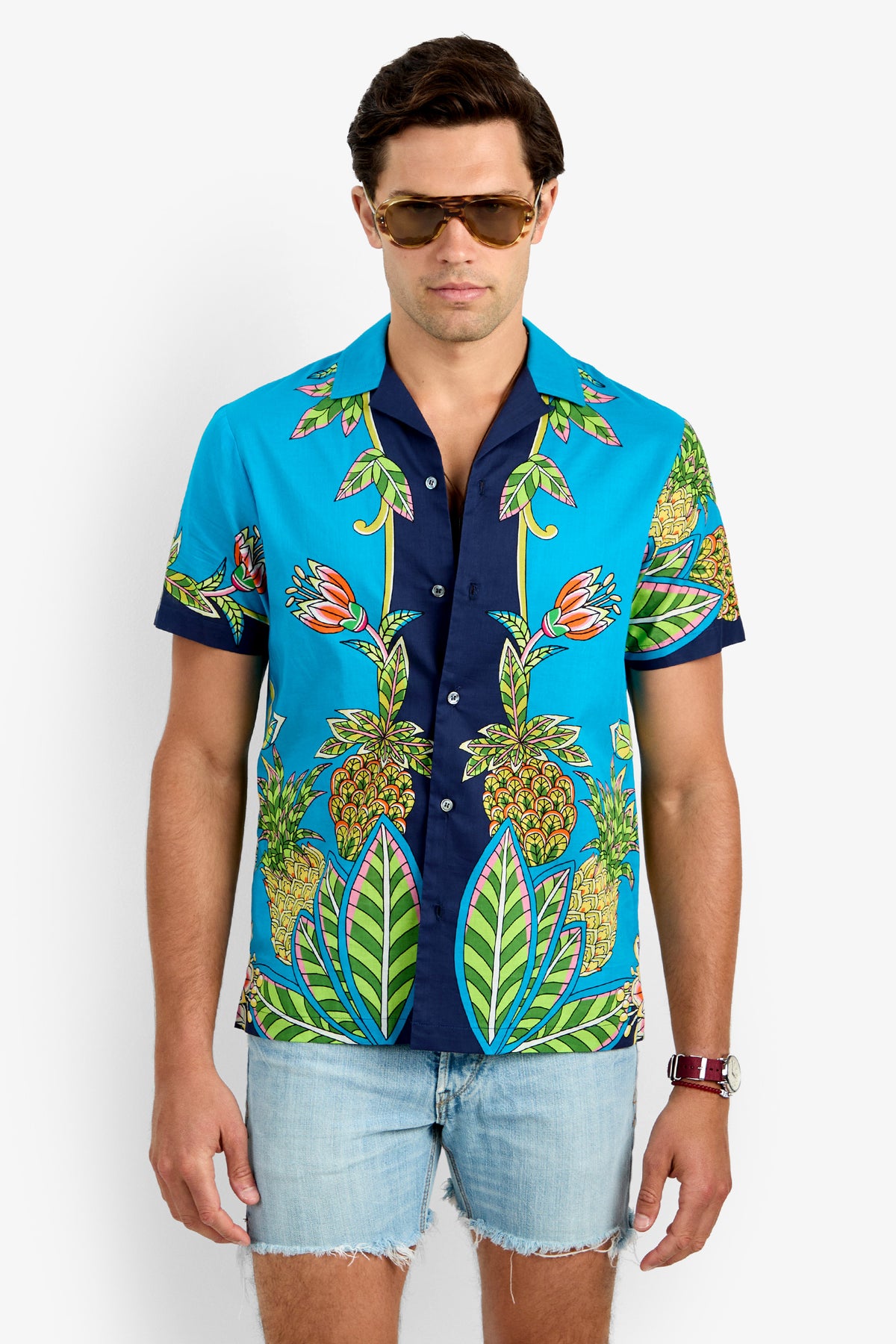 A man in sunglasses and light denim shorts wears the JOSUE CAMP SHIRT—a bright blue, 100% cotton, short-sleeve button-up with a colorful tropical print—standing against a plain white background.