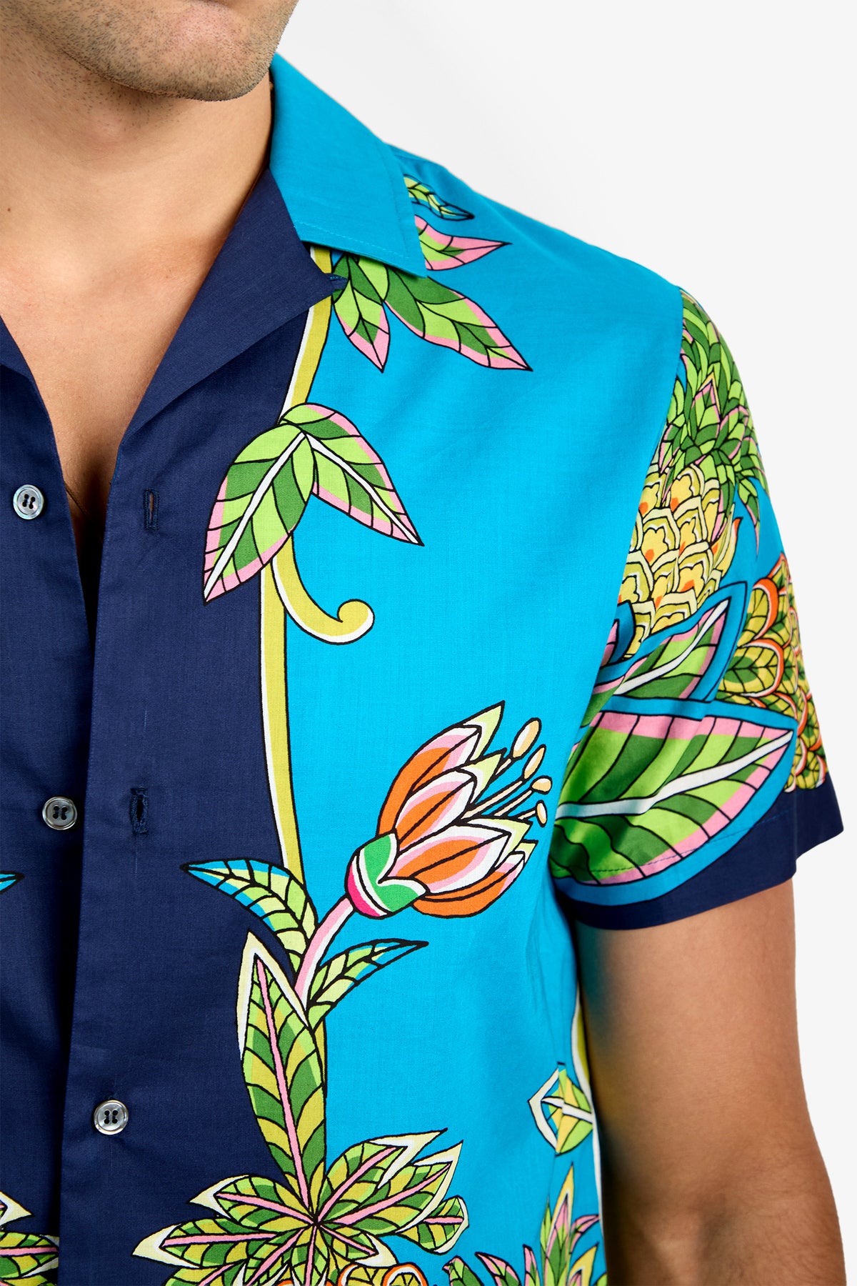 A close-up shows a person wearing the JOSUE CAMP SHIRT, a 100% cotton button-up featuring a vibrant floral and leaf pattern in blue, green, pink, and yellow. Only the lower face and upper torso are visible.