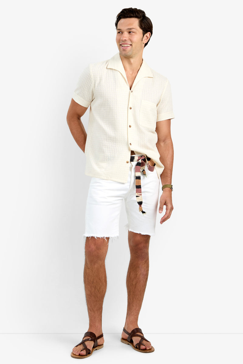 A man with short dark hair smiles against a light background, wearing THEO CAMP SHIRT in ECRU—a soft cotton short-sleeve button-up—paired with white frayed hem shorts, a striped belt, and brown sandals.