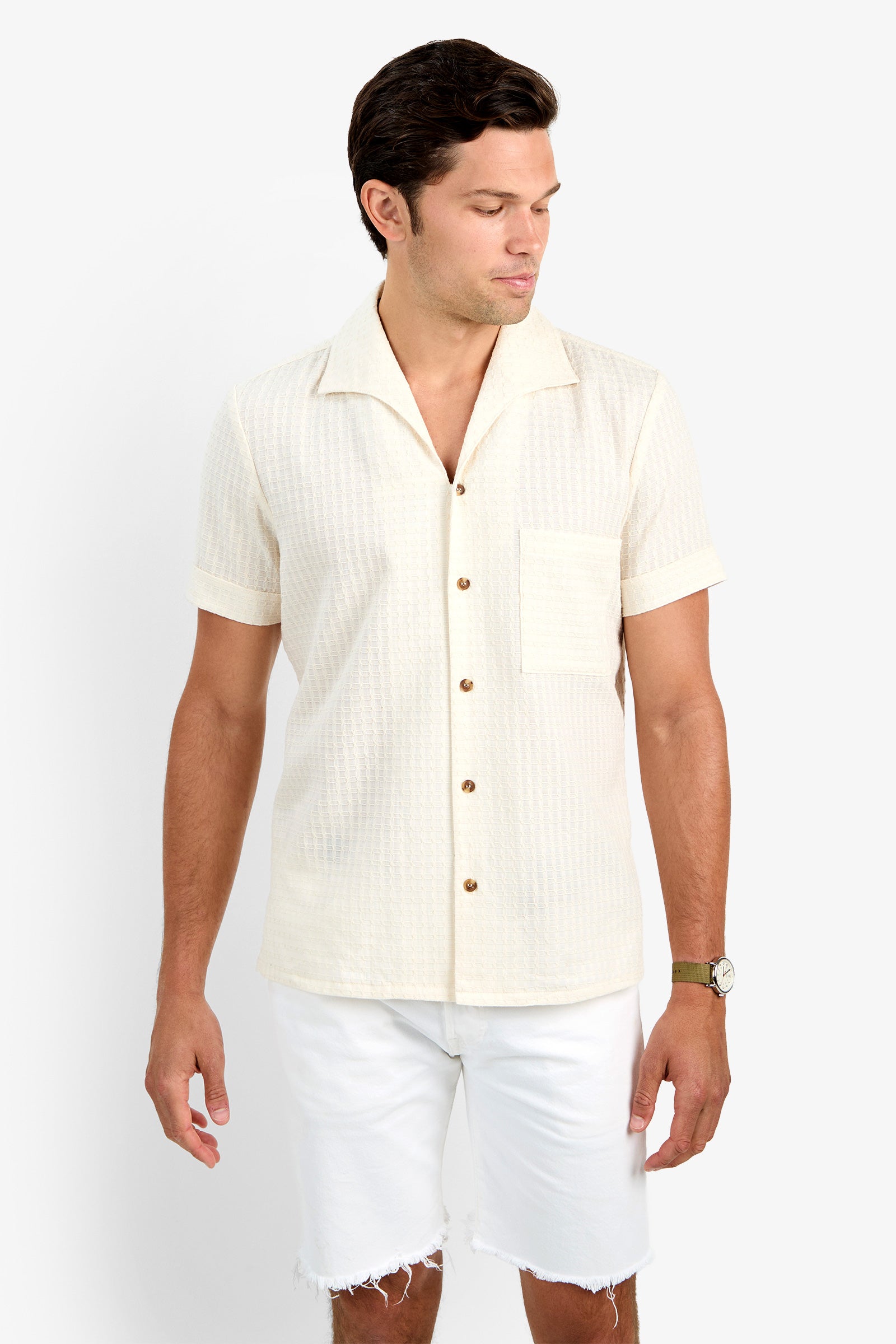 A man wearing the cream-colored THEO CAMP SHIRT with short sleeves, button-up front, soft cotton fabric, and a chest pocket stands against a white background, looking down and to the side while wearing white frayed-hem shorts.