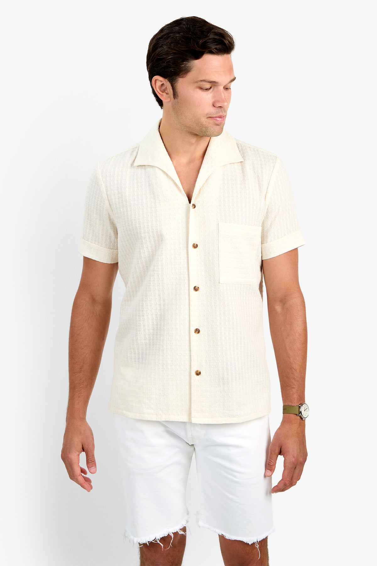 A man wearing the cream-colored THEO CAMP SHIRT with short sleeves, button-up front, soft cotton fabric, and a chest pocket stands against a white background, looking down and to the side while wearing white frayed-hem shorts.