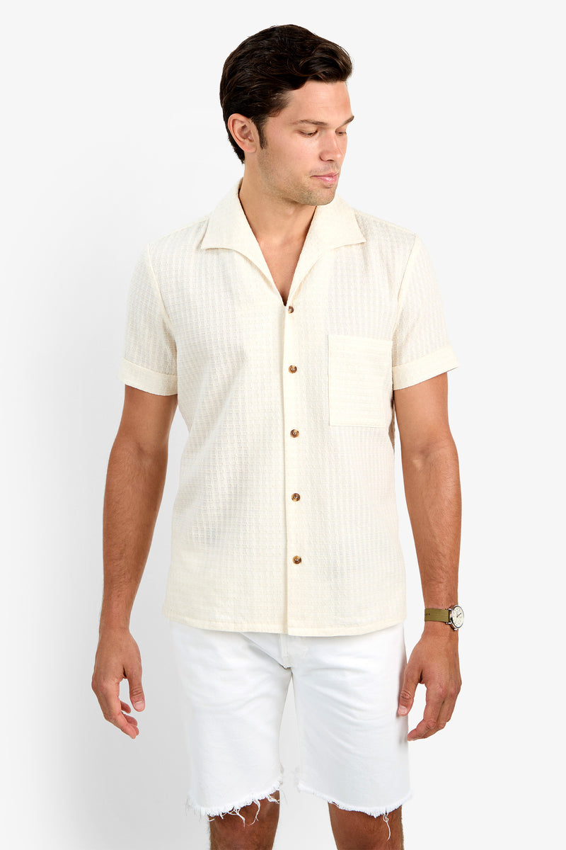 A man wearing the cream-colored THEO CAMP SHIRT with short sleeves, button-up front, soft cotton fabric, and a chest pocket stands against a white background, looking down and to the side while wearing white frayed-hem shorts.