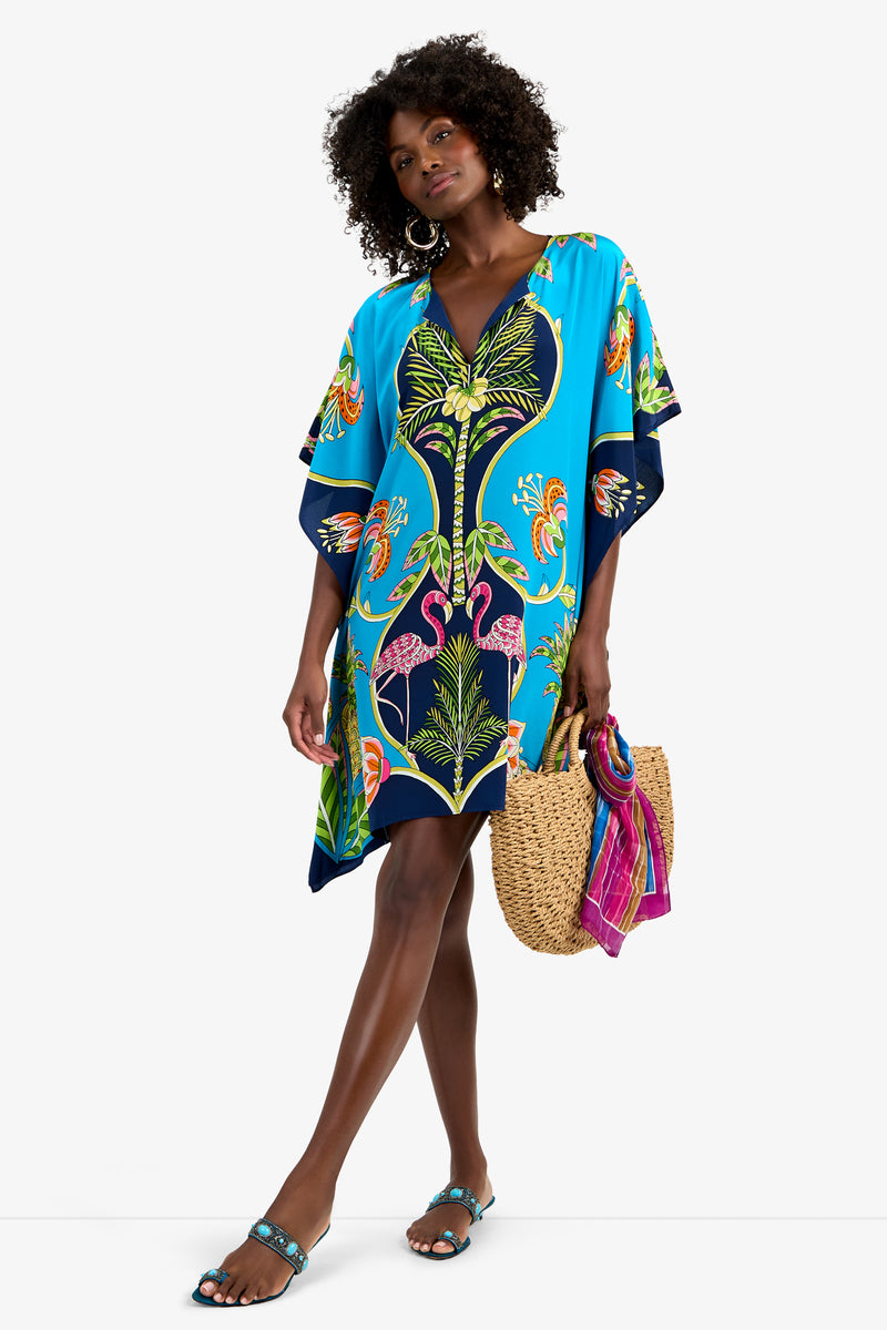 A woman with curly hair and large hoop earrings wears the THEODORA DRESS, a vibrant blue silk tropical print dress with flamingos. She carries a straw tote bag with a pink scarf and wears blue sandals.