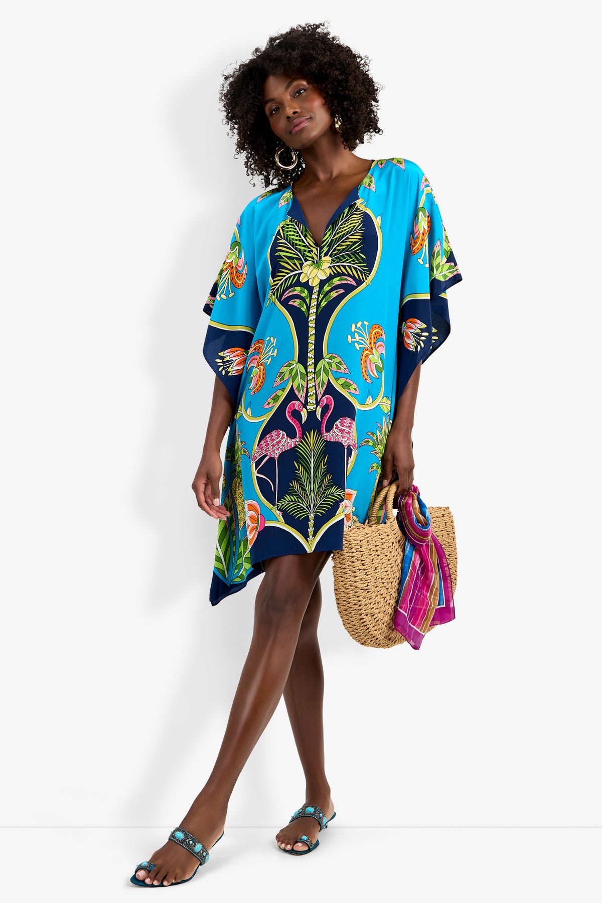 Woman in Theodora Caftan Dress with blue tropical print and pink flamingos, holding a straw bag with a pink scarf, wearing blue sandals and hoop earrings, standing against a white background.