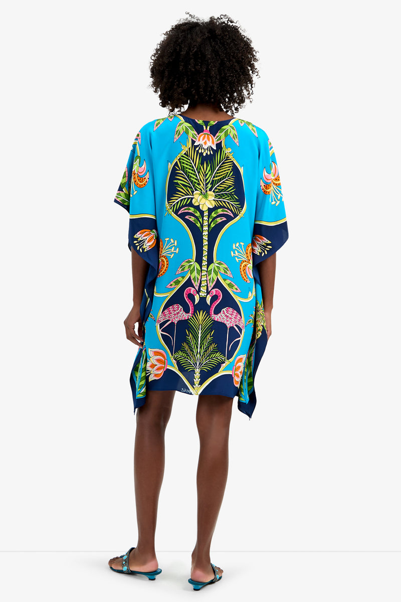 A woman with curly hair stands facing away, wearing THEODORA DRESS—a loose, brightly colored 100% silk kaftan featuring blue, green, orange, and pink tropical floral prints. She pairs it with blue sandals.
