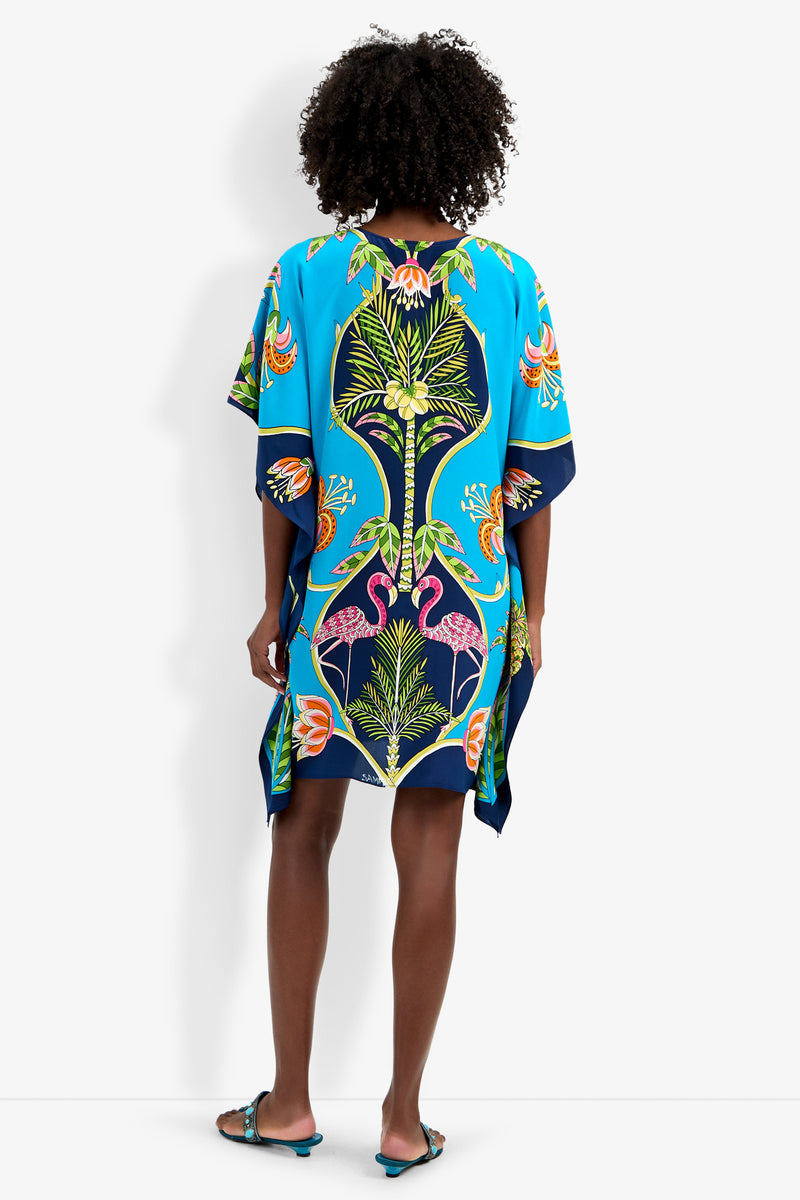 A person faces away, wearing Theodora Caftan Dress—a loose, vibrant blue piece with colorful botanical and floral patterns. The dress features wide sleeves, falls just above the knees, and is paired with flat sandals.