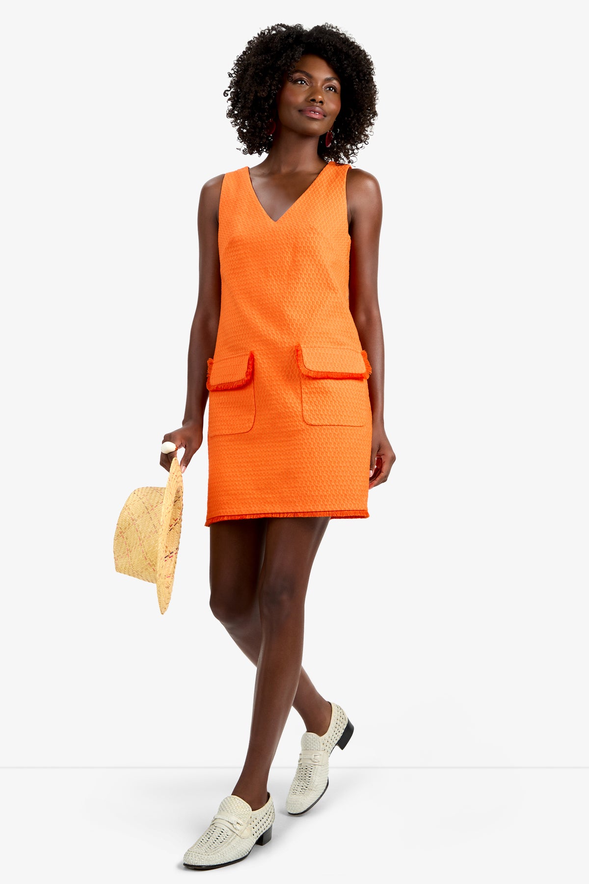 A woman with dark curly hair wears the WAVESON DRESS—a sleeveless, textured orange viscose dress with front pockets—paired with white slip-on shoes. She holds a straw hat and stands against a plain white background.