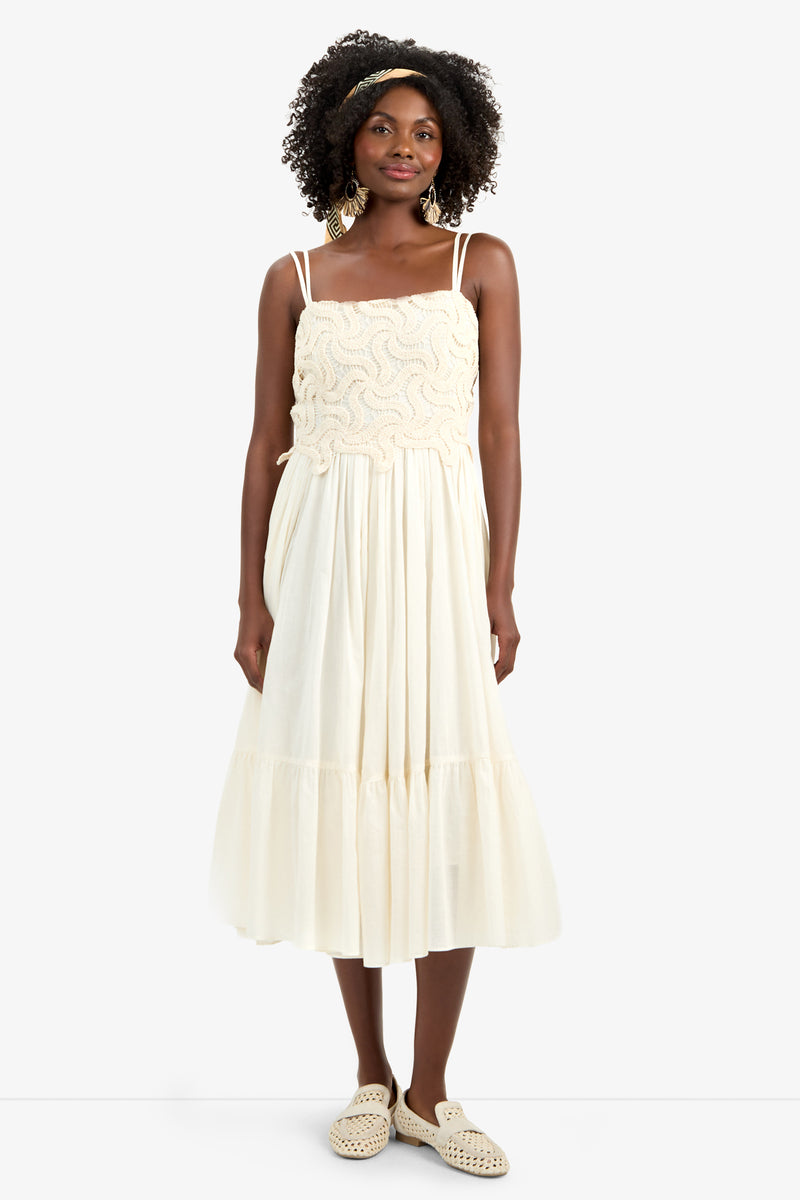 A woman with curly hair wears the BAYO DRESS, a cream-colored, sleeveless cotton dress with a textured bodice and flowing skirt, paired with woven slip-on shoes and gold earrings, standing against a plain white background.