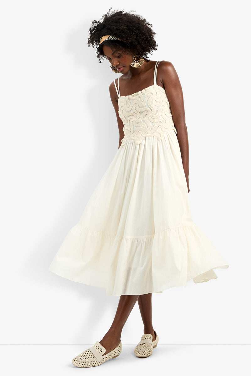 A woman wears the Bayo Dress, a sleeveless cream midi with a textured wavy bodice and tiered skirt. She styles it with curly hair, gold earrings, and woven cream loafers.