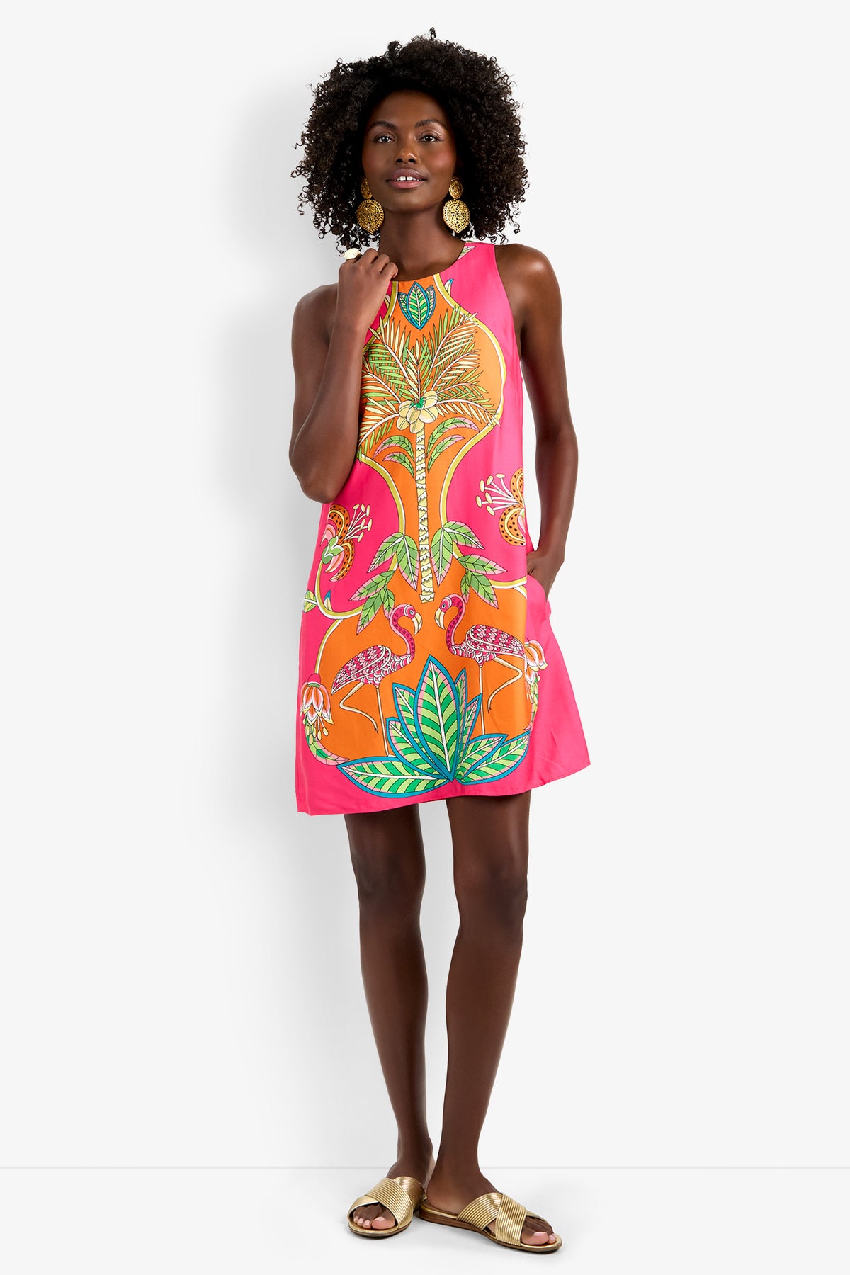A woman in the Loveage Dress, a sleeveless A-line with a vibrant tropical print, stands with one hand on her hip. She accessorizes with curly hair, gold earrings, and gold sandals against a plain white backdrop.