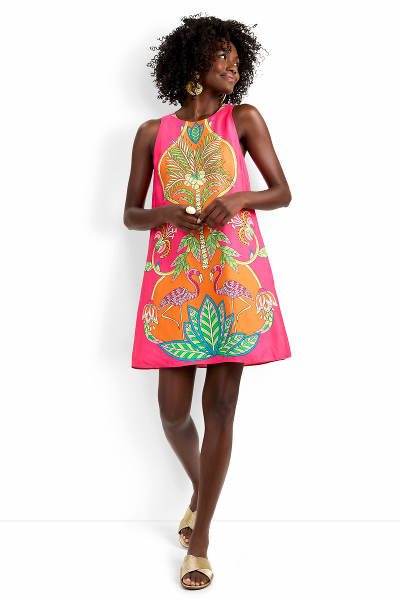 A woman models the Loveage Dress, a vibrant pink sleeveless piece adorned with bold tropical flamingo, leaf, and flower designs. She pairs it with large earrings and beige sandals, posing against a plain white background.