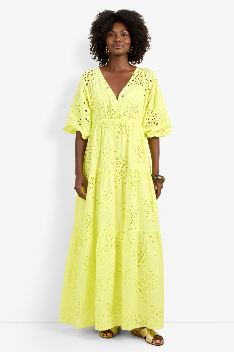 A woman with curly hair stands against a white background wearing the Arco Iris Dress, a long, bright yellow eyelet gown with puffed sleeves and a v-neckline. She pairs it with sandals and bracelets.