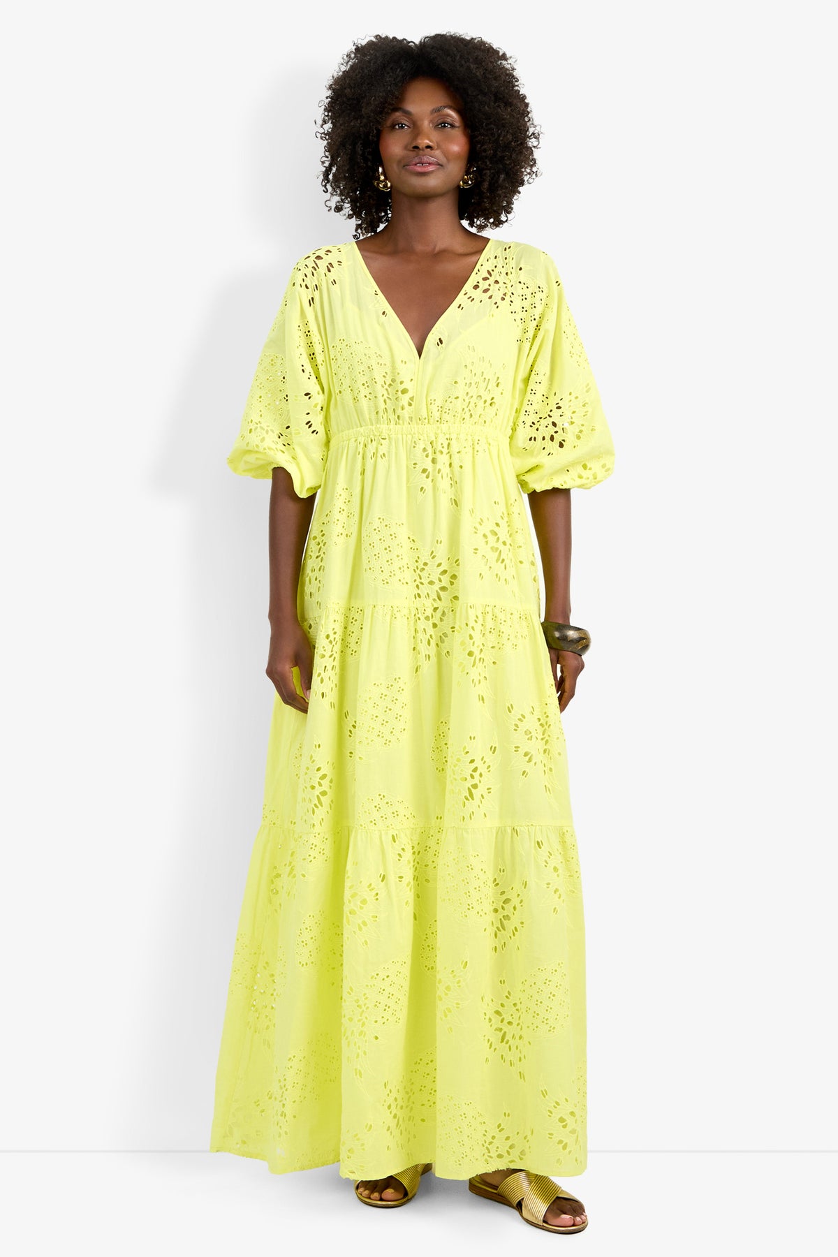 A woman with curly hair wears the Arco Iris Dress, a long yellow eyelet maxi featuring a V-neck, tiered skirt, and puff sleeves. She accessorizes with bracelets and flat sandals.