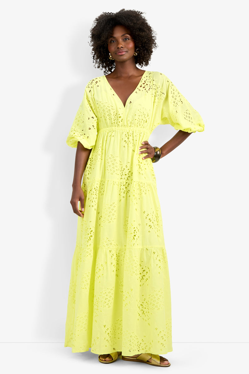 A woman with curly hair, gold earrings, and a bracelet stands facing forward in the Arco Iris Dress—a bright yellow, eyelet-patterned maxi dress with puffed sleeves—paired with sandals.