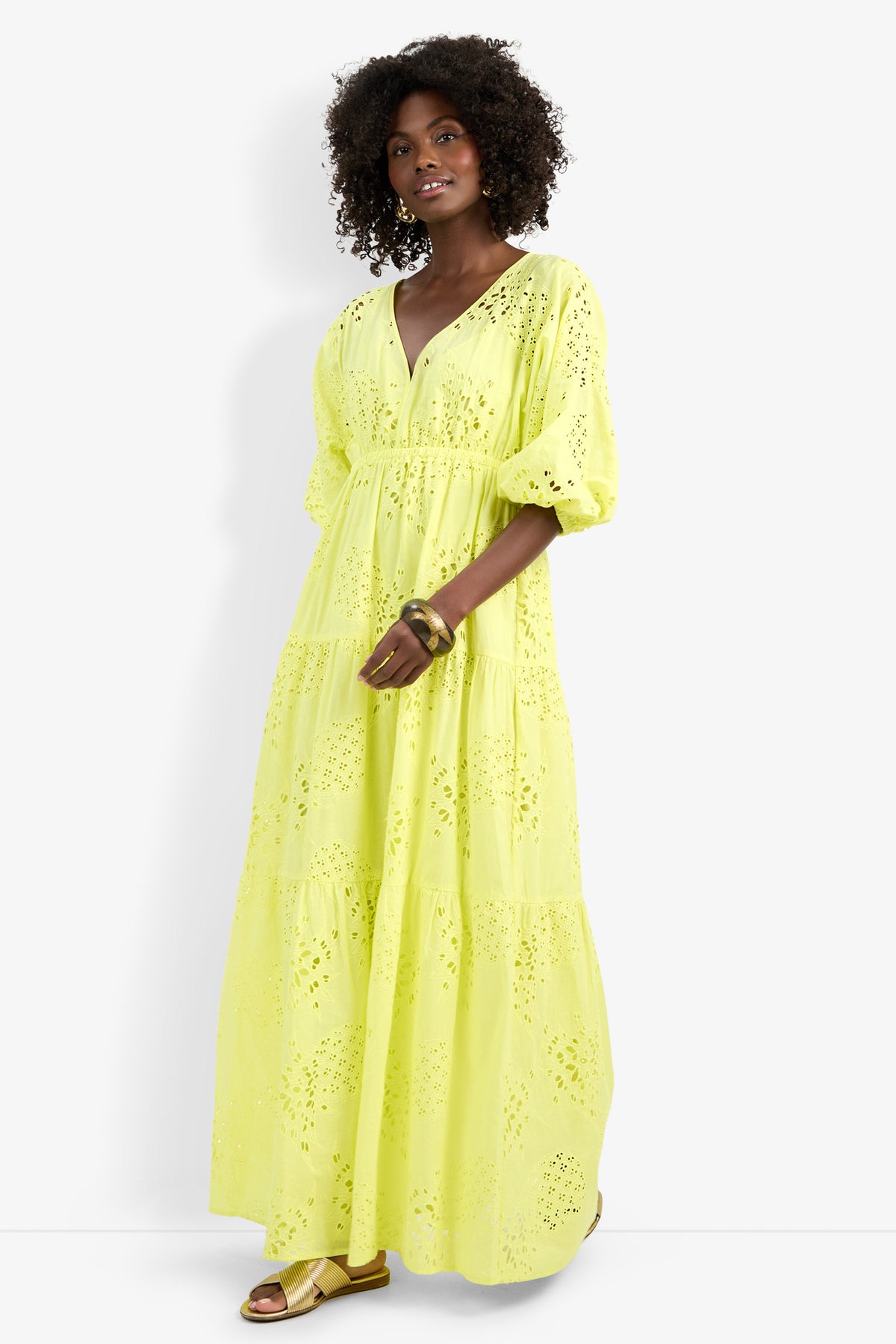 A woman with curly hair wears the Arco Iris Dress—a long, bright yellow eyelet dress with puffed sleeves and a V-neck—paired with gold sandals and bangles, standing against a plain white background.