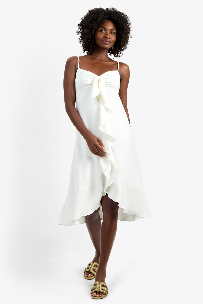 A woman models the LILLA DRESS—a sleeveless white dress with spaghetti straps, front ruffle detail, and lightweight polyester fabric—paired with gold sandals against a plain white background.