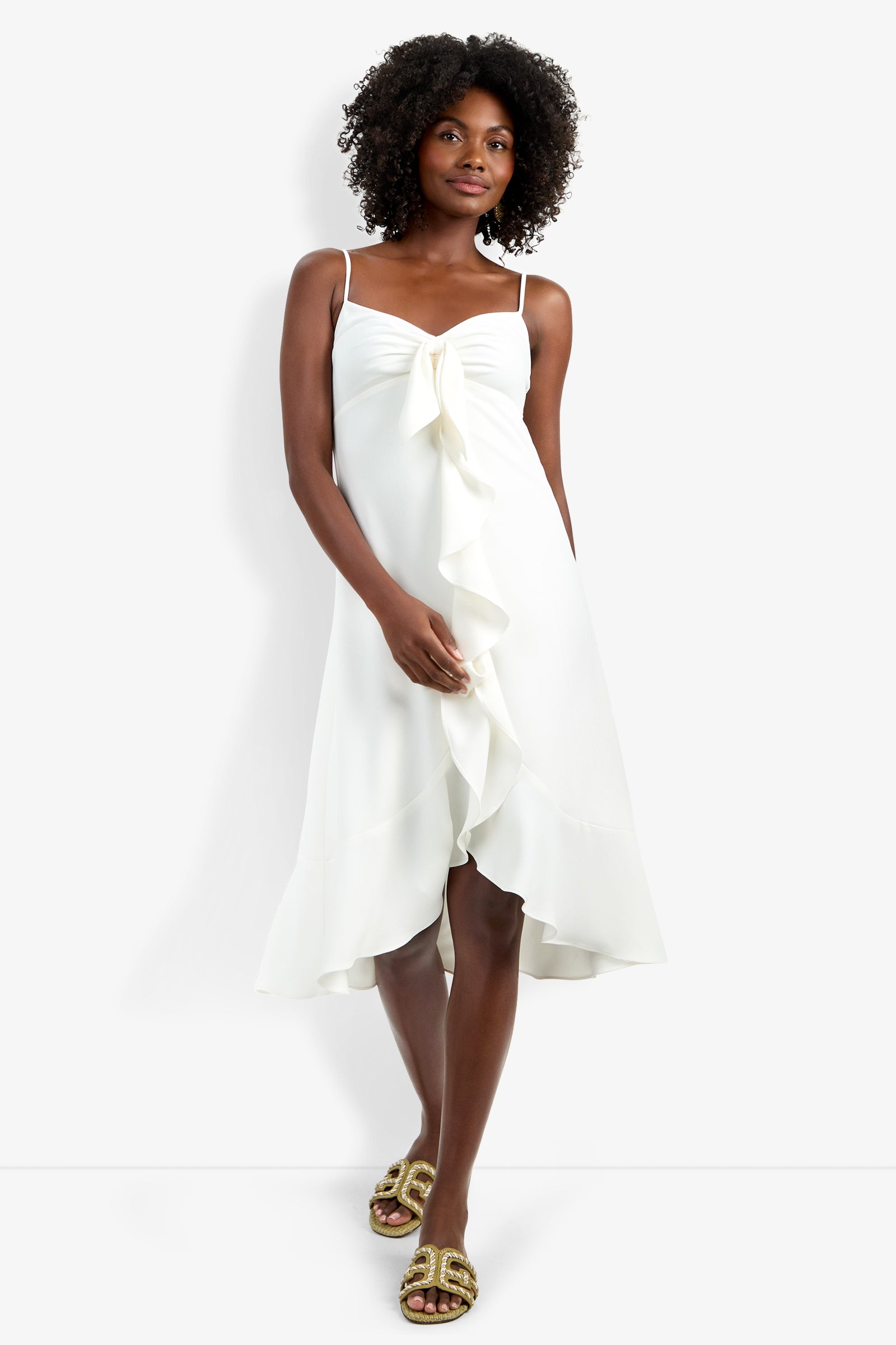 A woman models the sleeveless, knee-length Lilla Dress in white, featuring a front ruffle and bow detail, paired with gold sandals against a plain white background.