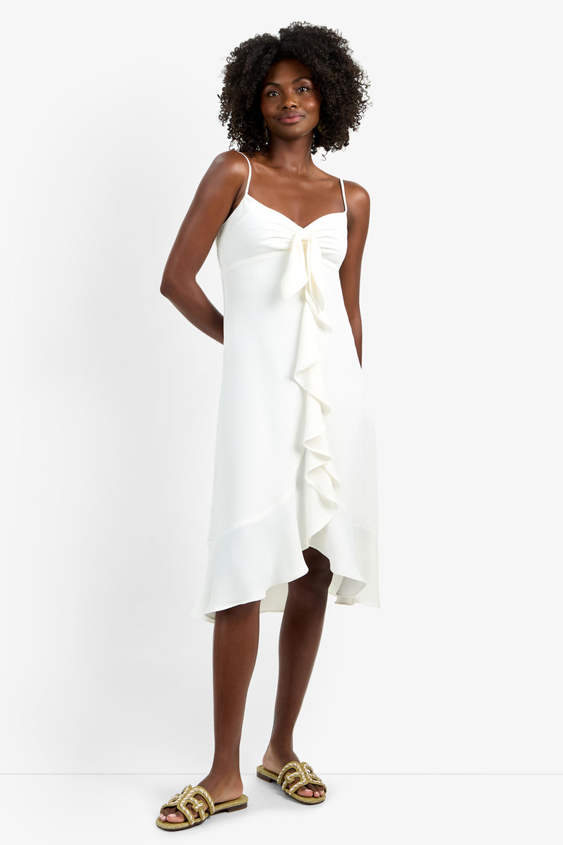 A woman with short curly hair wears the LILLA DRESS—a white sleeveless, lightweight polyester dress with a front tie and ruffled asymmetrical hem—paired with gold slide sandals, standing against a plain white background.