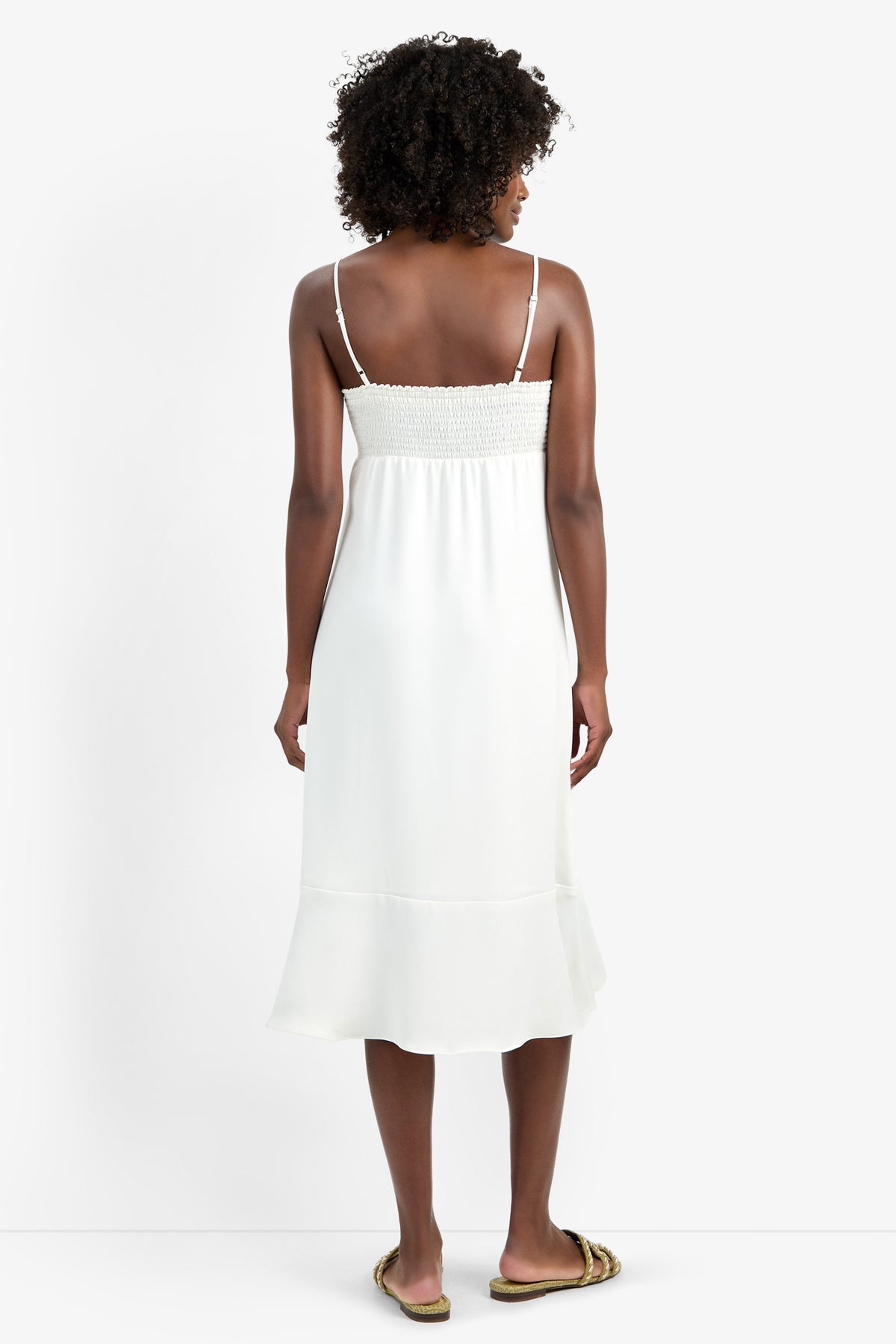 A woman with curly hair, seen from behind, wears the LILLA DRESS—a white, sleeveless, mid-length dress with thin straps and a ruffled hem—paired with flat sandals, standing against a plain white background.