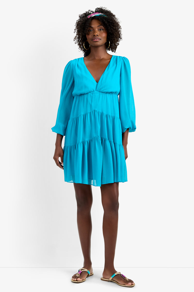 A woman with curly hair and hoop earrings wears the MAKE MERRY DRESS, a bright blue, long-sleeve, V-neck tiered mini dress in soft polyester with gathered details. She pairs it with sandals and a matching blue headband against a white background.