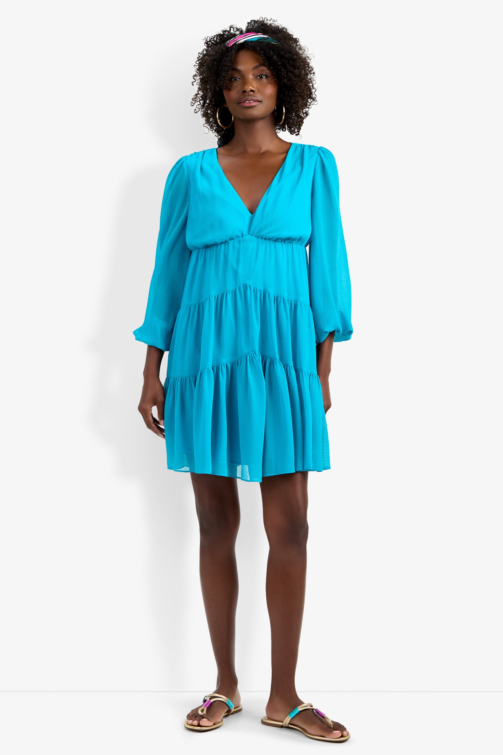 A woman wearing the Make Merry Dress—a bright blue, long-sleeve, V-neck, tiered mini dress—paired with hoop earrings, a blue headband, and decorative flat sandals stands against a plain white background.