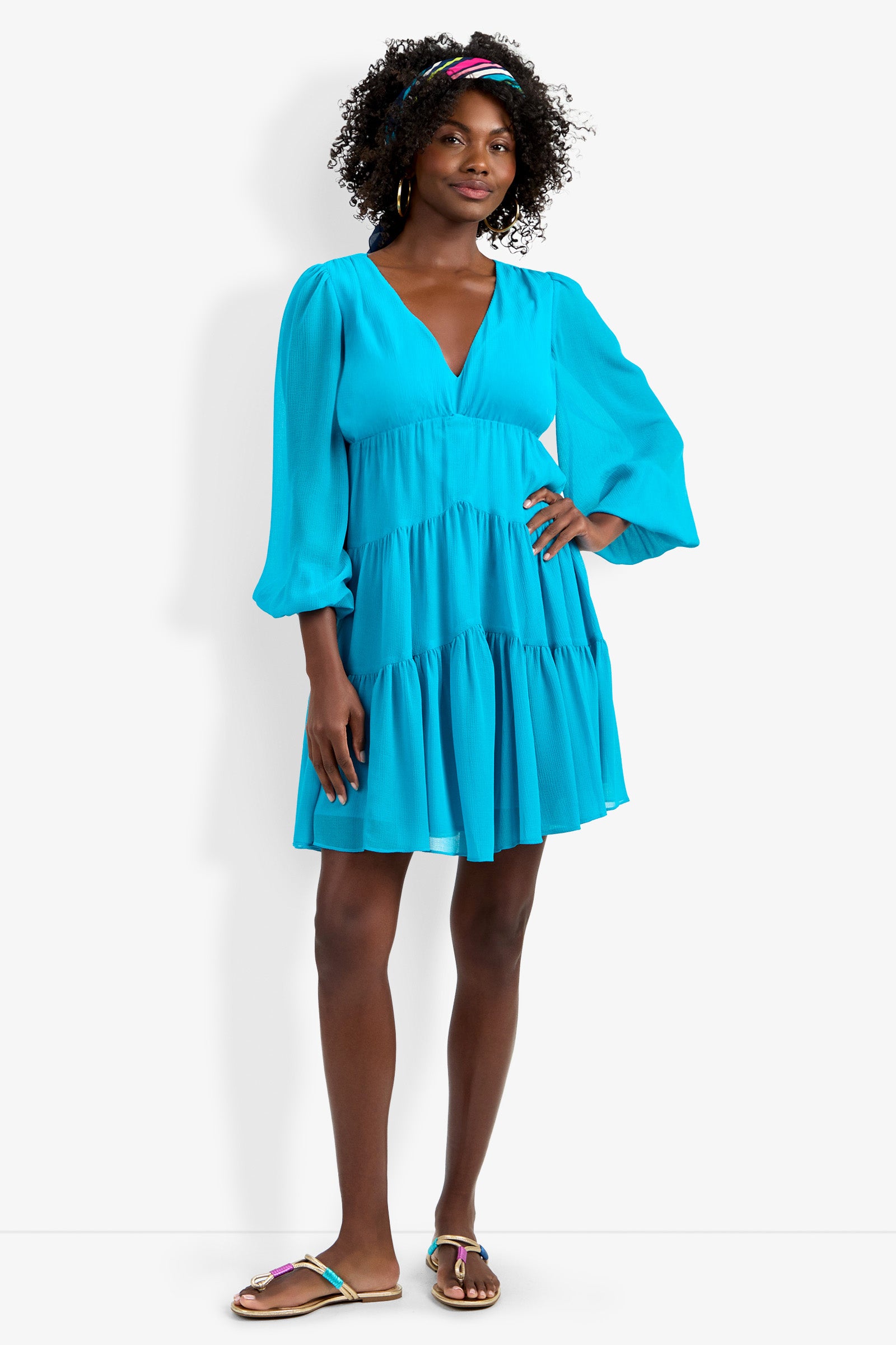 A woman with curly hair in a colorful headband smiles, wearing the Make Merry Dress—a bright blue, long-sleeve, tiered V-neck dress with puff sleeves—and sandals against a plain white background.