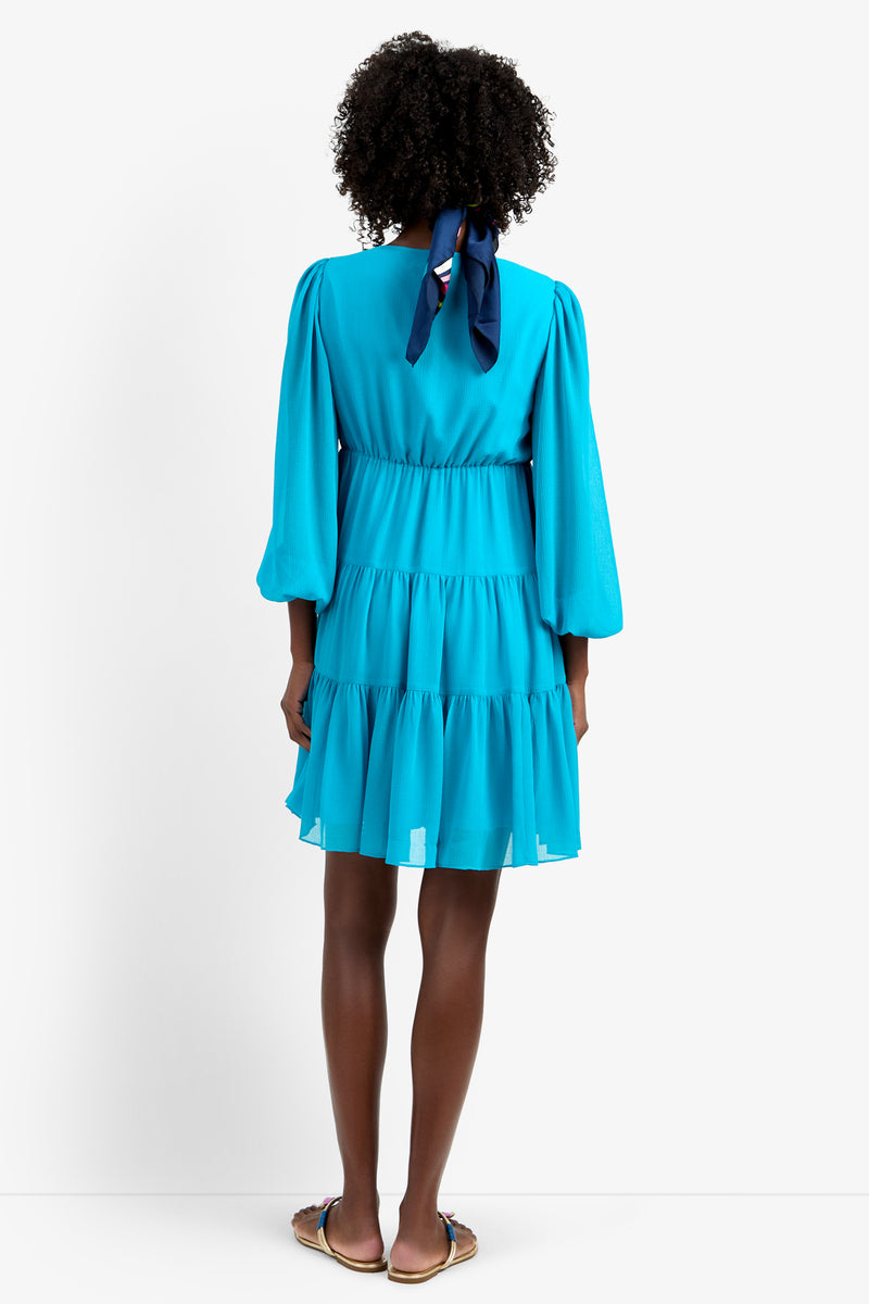 A woman with a navy ribbon in her curly hair wears the MAKE MERRY DRESS, a bright blue, long-sleeve, tiered dress made from 100% polyester. She completes the look with flat sandals against a plain white background.