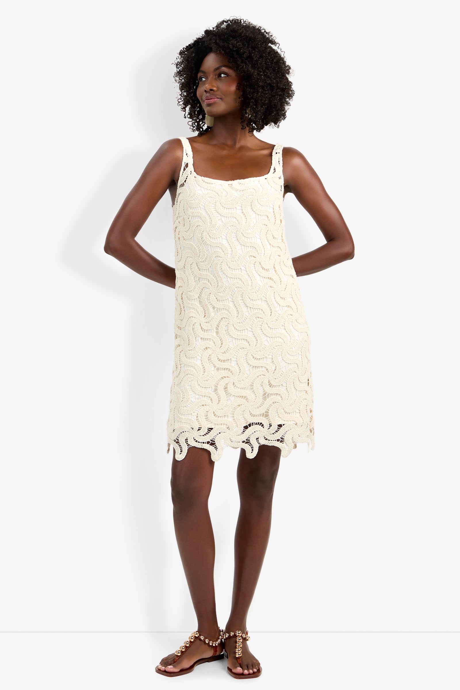 A woman wearing the Aspire Dress, a sleeveless cream crochet dress with a wavy pattern and scalloped hem, stands with her hands on her hips and brown sandals.