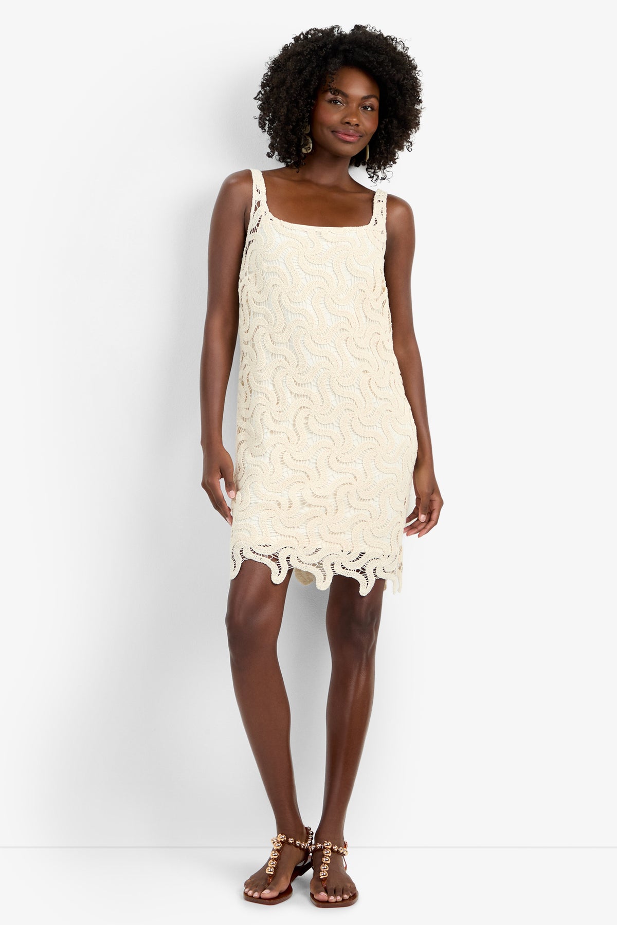 A woman with curly hair wears the ASPIRE DRESS, a cream, sleeveless crochet mini dress in 100% cotton featuring a wavy pattern and scalloped hem, paired with studded flat sandals against a white background.