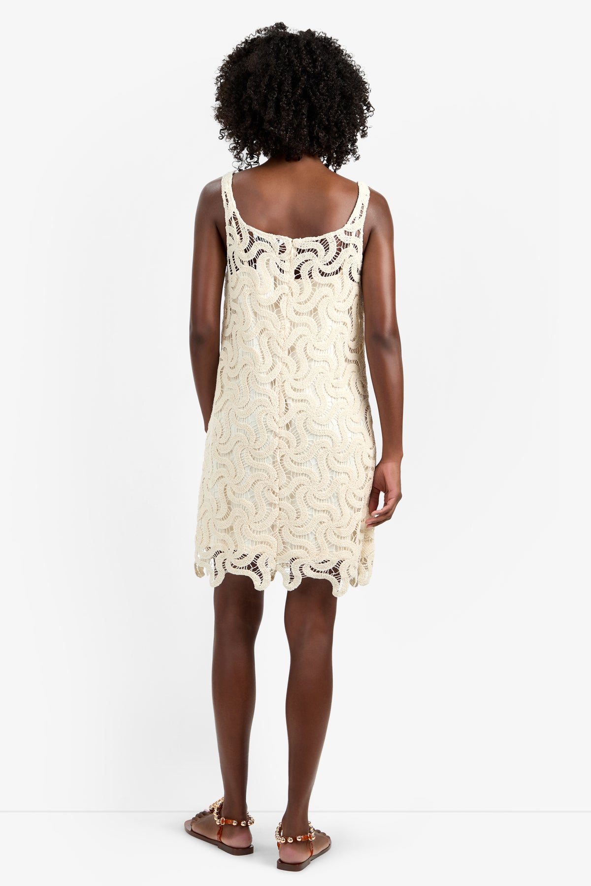 A woman with curly hair stands facing away, wearing the ASPIRE DRESS—a sleeveless, cream crochet dress in 100% cotton with a wavy pattern and scalloped hem—paired with brown flat sandals, against a plain white background.