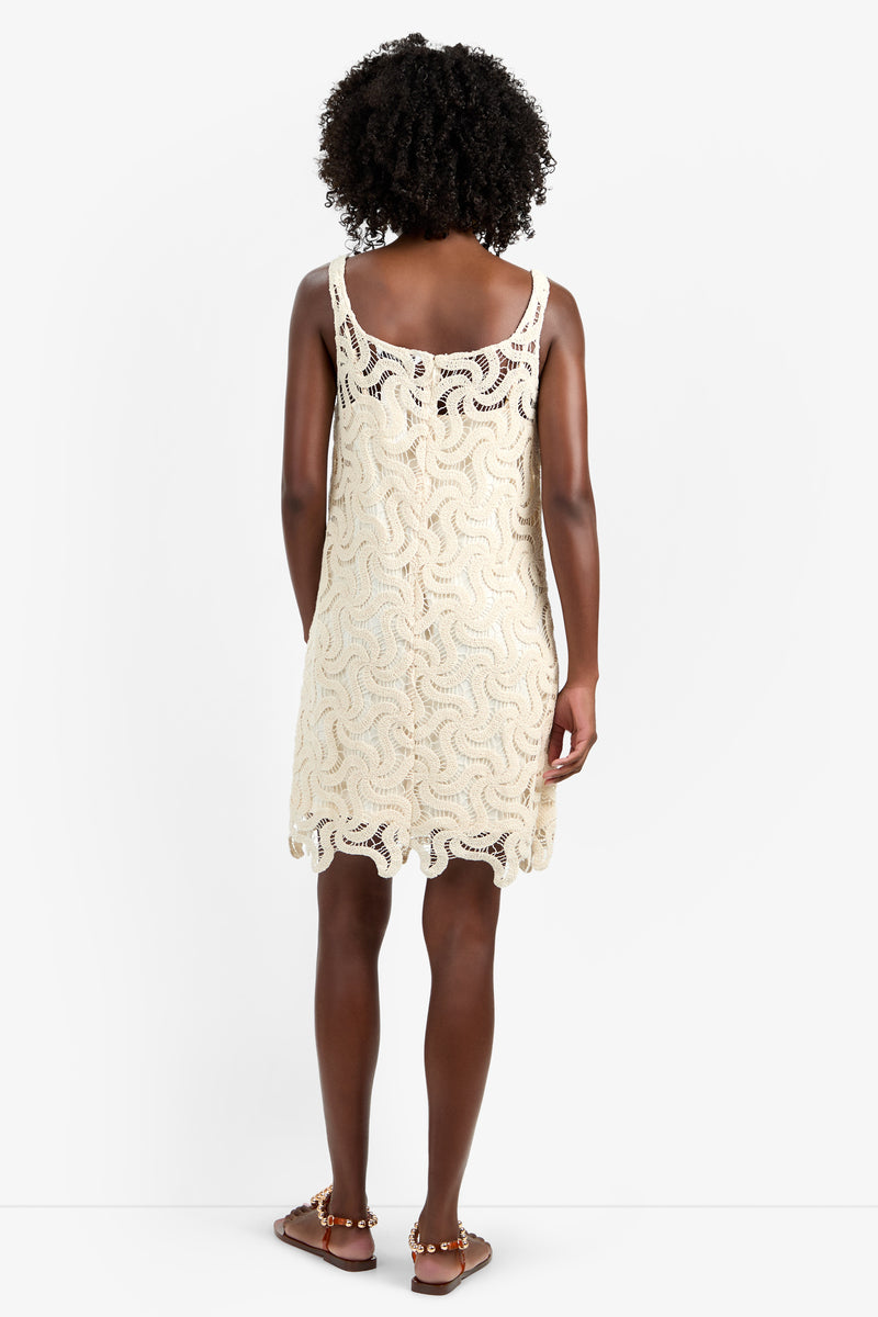 A woman with curly hair stands facing away, wearing the ASPIRE DRESS—a sleeveless, cream crochet dress in 100% cotton with a wavy pattern and scalloped hem—paired with brown flat sandals, against a plain white background.