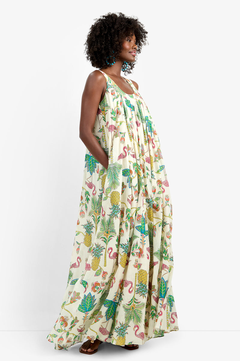 A woman stands sideways with her hand in her pocket, wearing the JIANNA MAXI DRESS—a long 100% cotton dress featuring a colorful tropical print with pineapples, flamingos, and leaves—against a plain white background.