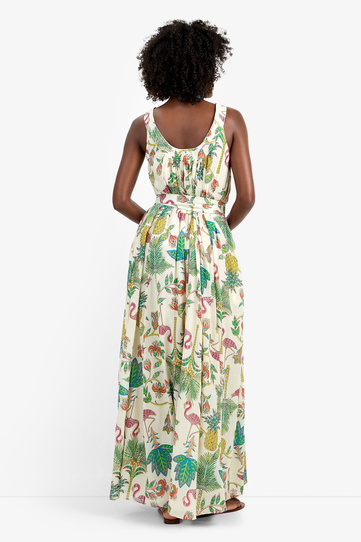 A woman with curly hair stands against a white background, wearing the JIANNA MAXI DRESS—a sleeveless, floor-length 100% cotton dress featuring a vibrant tropical print of leaves, flamingos, and pineapples.