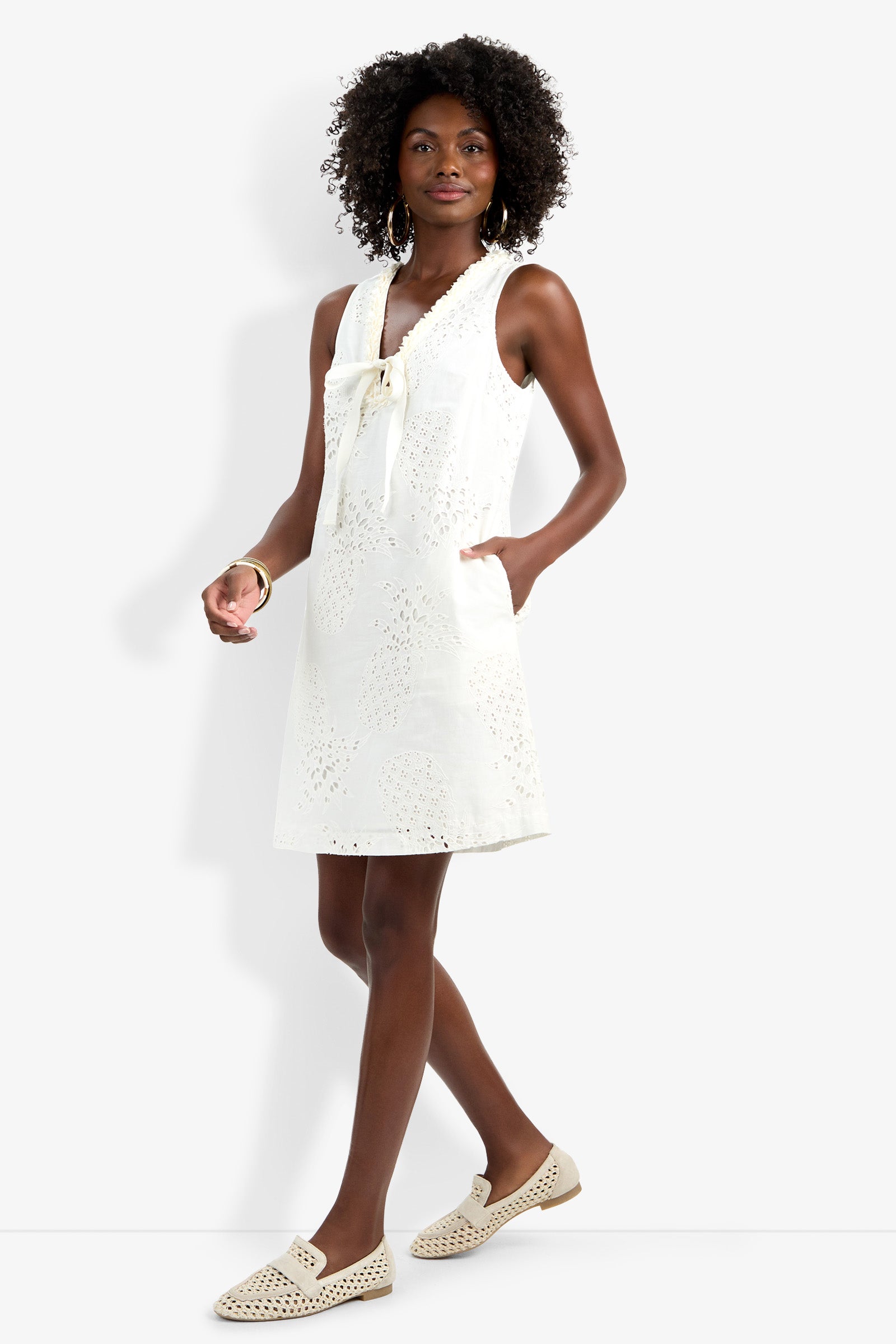 A woman stands confidently in the Amala Dress—a sleeveless, knee-length white dress with eyelet details and a front tie—paired with beige slip-on shoes and hoop earrings against a plain light background.