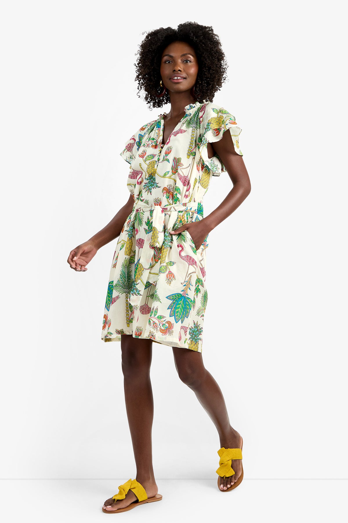 A woman smiles while walking in the ZANDY DRESS—a short, white 100% cotton dress with colorful tropical prints, ruffled sleeves, and a tied waist—paired with yellow bow sandals against a plain white background.