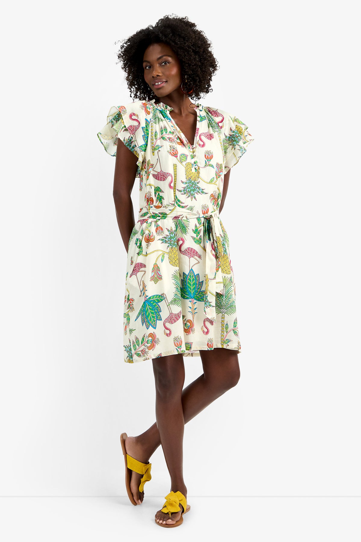 A woman with curly hair poses with one leg crossed, wearing the ZANDY DRESS in MULTI—a short-sleeve, 100% cotton white dress featuring a colorful tropical print, tie waist, and ruffled sleeves—paired with yellow sandals.
