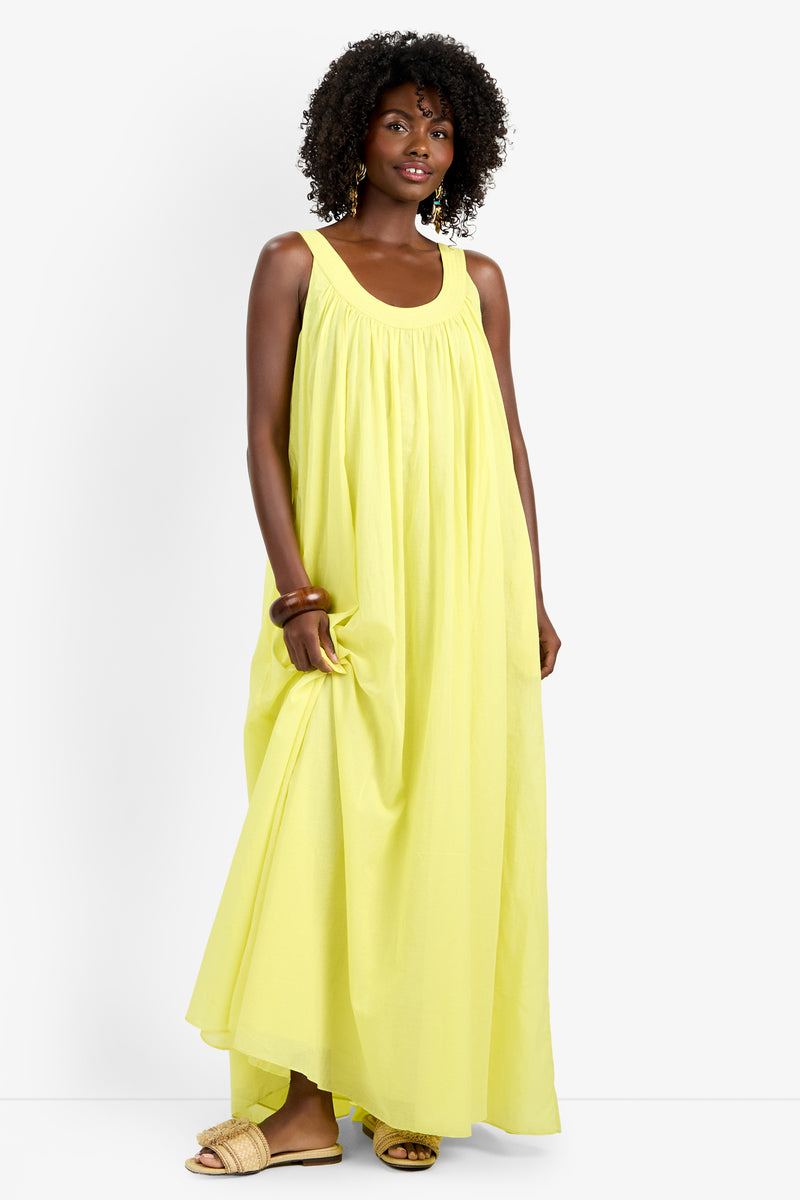 A woman with curly hair wears the JIANNA DRESS, a flowing, sleeveless yellow 100% cotton maxi, and beige slip-on sandals. Against a plain white background, she holds part of the dress and smiles softly.