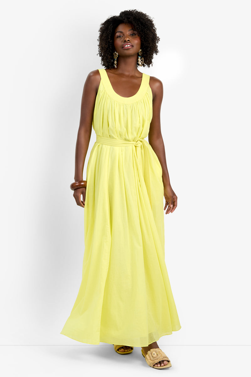 A woman wearing the sleeveless, yellow JIANNA DRESS with a tied waist stands against a plain white background, styled with gold statement earrings, bangles, and woven sandals.
