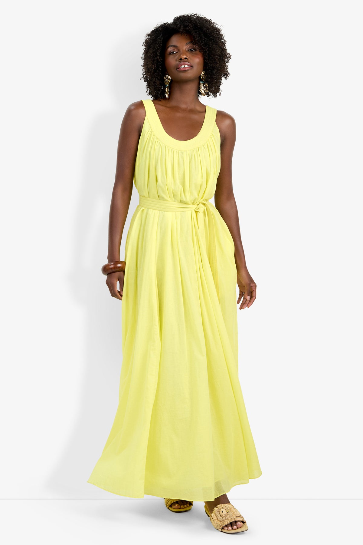 A woman with curly hair and gold earrings poses confidently against a white background in the sleeveless, light yellow Jianna Dress featuring a belted waist, styled with sandals.
