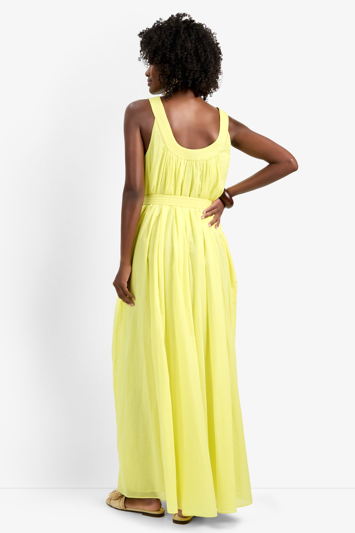 A woman with curly hair stands with her back to the camera, modeling the JIANNA DRESS—a sleeveless, floor-length light yellow dress in 100% cotton, featuring a gathered waist and flowing skirt. She pairs it with flat sandals.