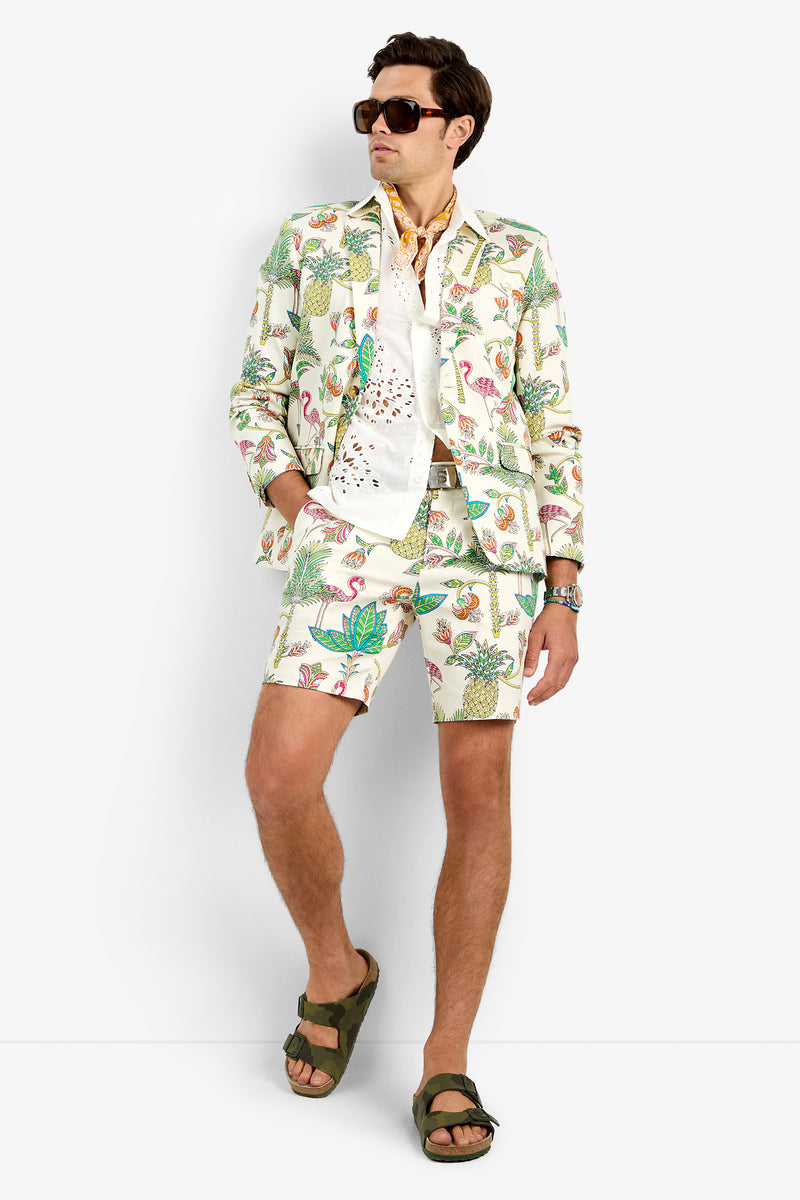 A man wearing the THURSTON BLAZER—a white cotton tropical print—matching shorts, a white shirt, sunglasses, and green sandals stands with one hand in his pocket, gazing to the side against a plain white background.