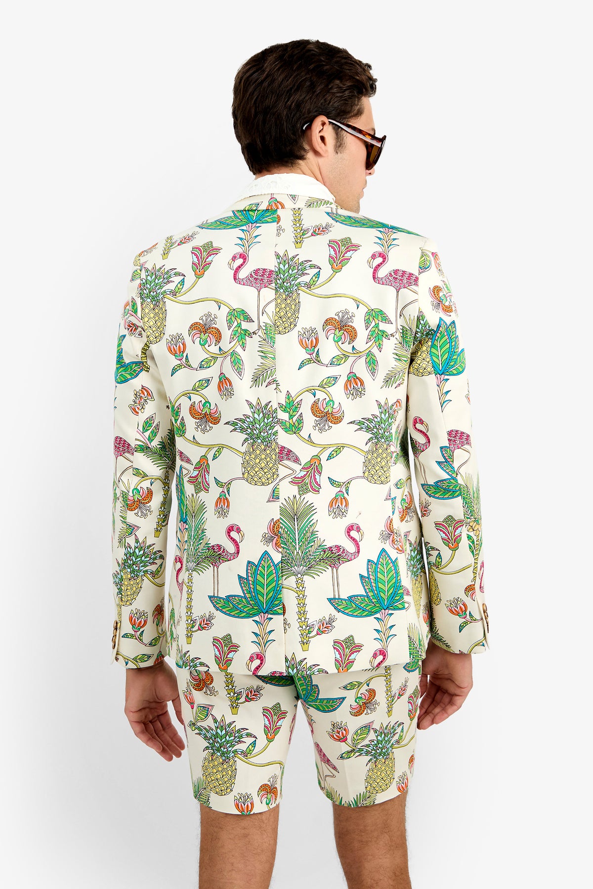 A man, seen from behind against a plain white background, wears the THURSTON BLAZER and matching shorts—white cotton with a colorful tropical print of pineapples, flamingos, and leaves—paired with sunglasses.