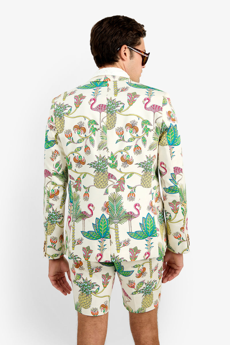 A man, seen from behind against a plain white background, wears the THURSTON BLAZER and matching shorts—white cotton with a colorful tropical print of pineapples, flamingos, and leaves—paired with sunglasses.