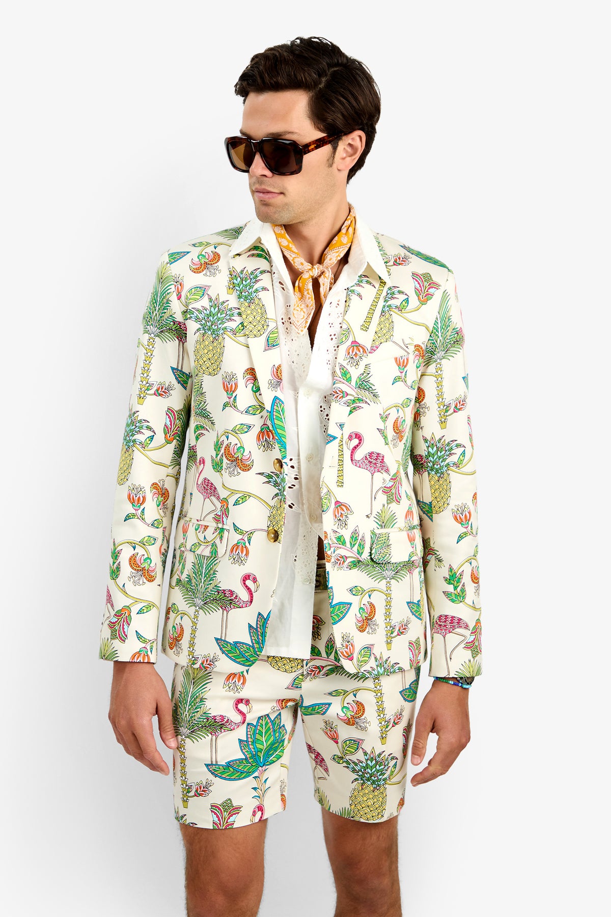 A man wearing sunglasses and the THURSTON BLAZER with matching shorts, featuring colorful tropical prints of flamingos, pineapples, and leaves, stands against a plain white background.